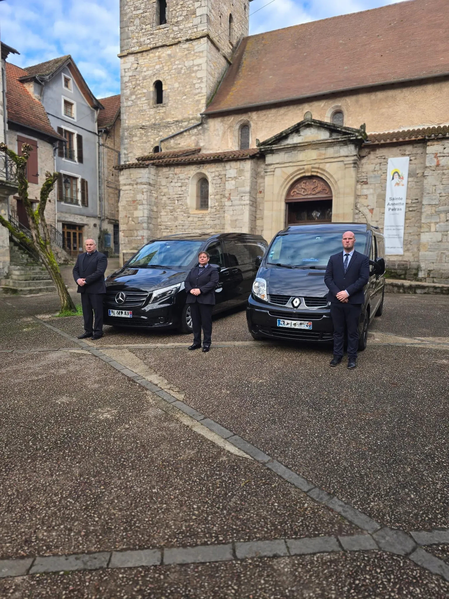 Trois agents funéraires en costume devant deux corbillards et une église.